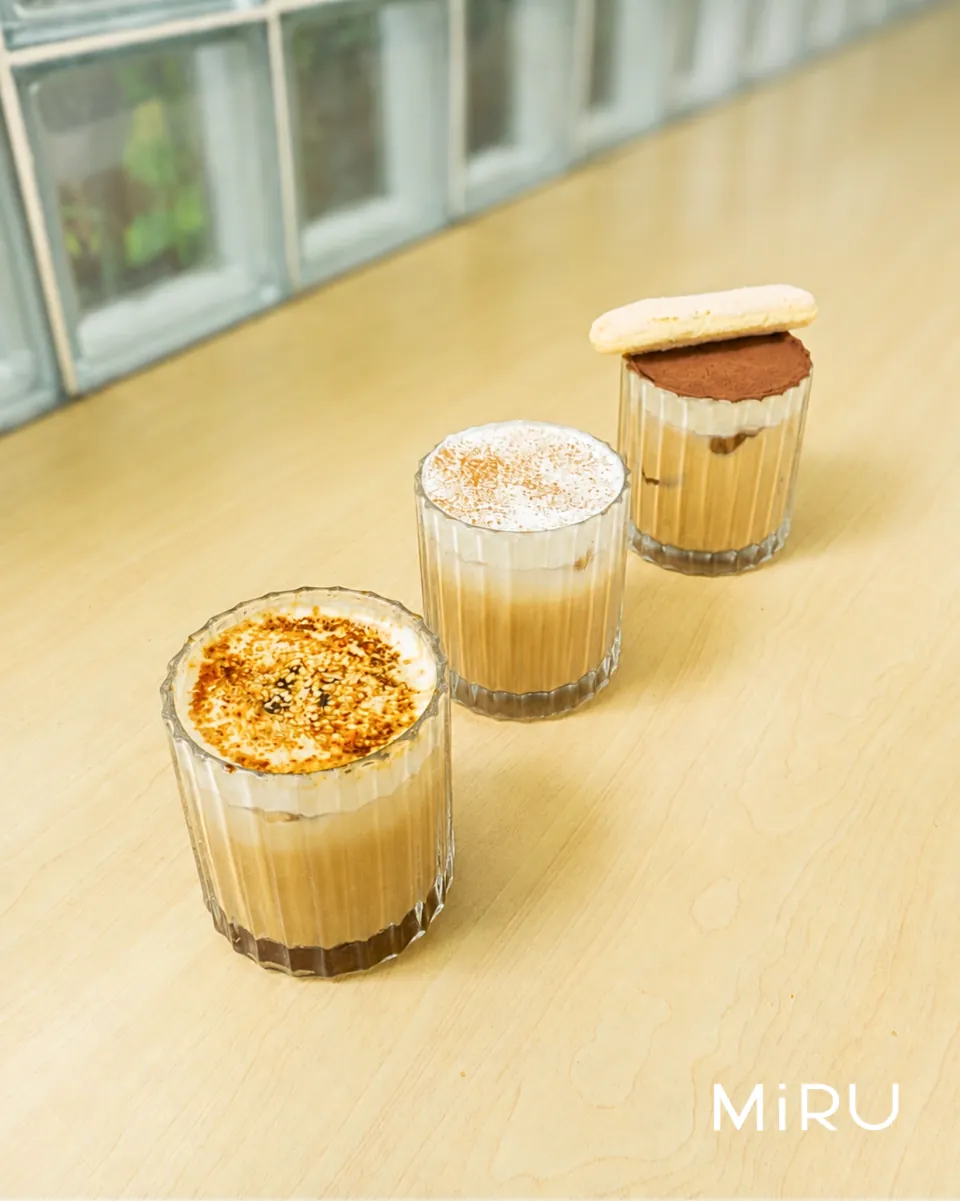 Tiga minuman kopi MiRU dalam gelas bergaris di atas meja kayu terang, dengan wordmark MiRU